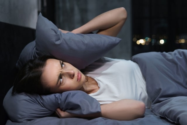 Frau liegt im Bett, hält sich Kissen über Ohren, Lärm von Nachbarn